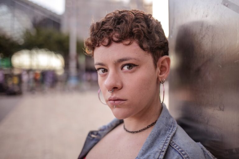 Close-up of a person with short curly brown hair, nose ring, lip piercing, and hoop earrings, wearing a denim jacket, leaning against a metal wall.