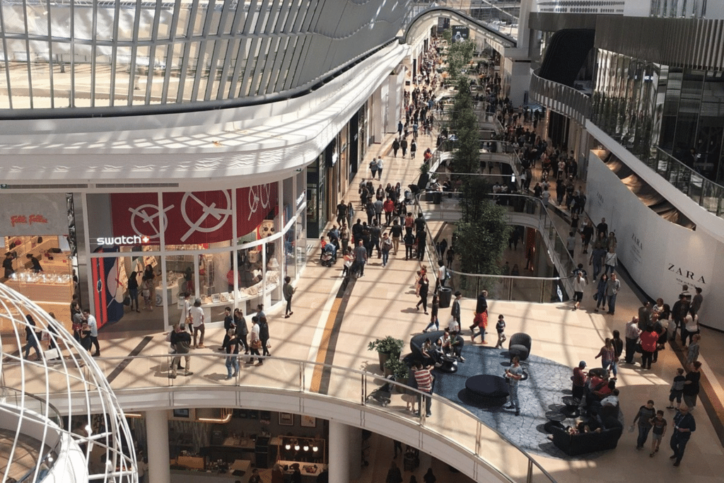 A bustling indoor shopping mall filled with shoppers walking and browsing various stores.