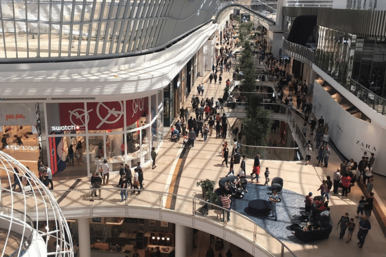 A bustling indoor shopping mall filled with shoppers walking and browsing various stores.