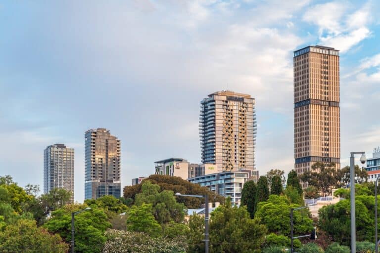 A city skyline featuring tall buildings alongside lush green trees under a clear blue sky.
