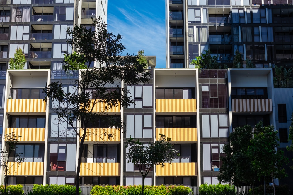The image shows a modern apartment building with multiple floors. The facade features a combination of glass windows and yellow-striped panels. There are trees and greenery in front of the building, and the sky is clear with some wispy clouds.