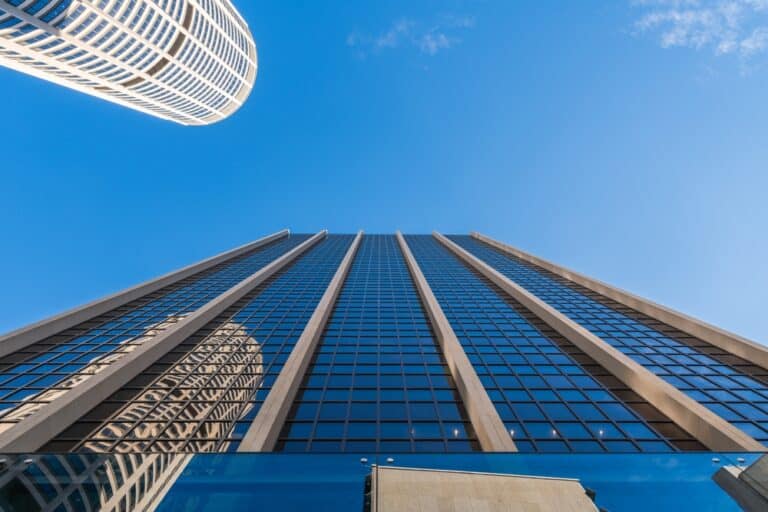 The image shows an upward view of a tall, modern skyscraper with a glass facade reflecting the blue sky. Another cylindrical building is visible on the left side of the image, also reflecting the sky. The perspective emphasizes the height and architectural design of both buildings against a clear blue sky with minimal clouds.