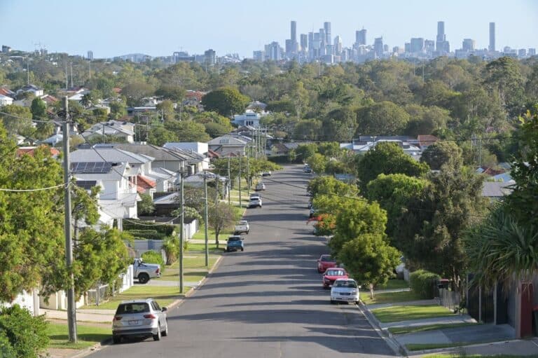 A street lined with trees, featuring houses in the background under a clear blue sky.