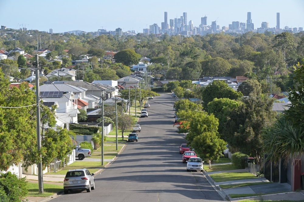 A street lined with trees, featuring houses in the background under a clear blue sky.