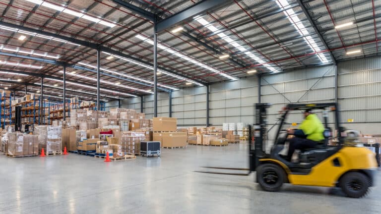 The image shows a large warehouse with stacks of boxes and pallets. A person in a high-visibility jacket is driving a yellow forklift across the concrete floor.