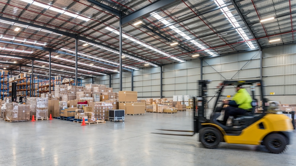 The image shows a large warehouse with stacks of boxes and pallets. A person in a high-visibility jacket is driving a yellow forklift across the concrete floor.