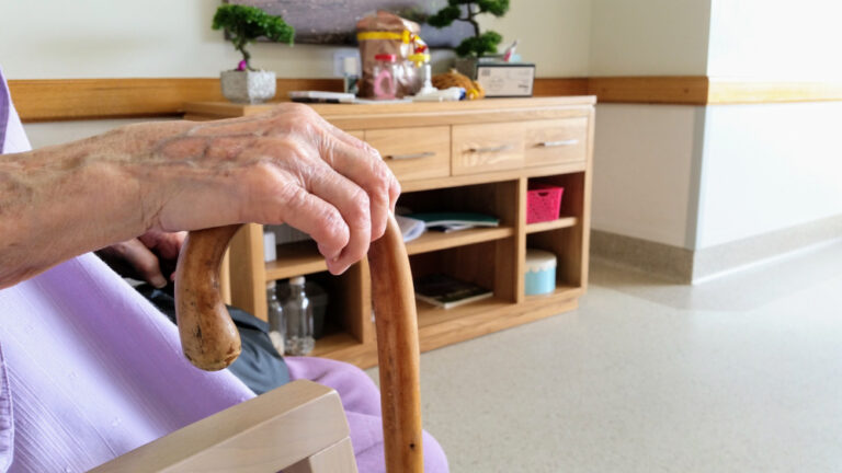 The image shows an elderly person's hand resting on a wooden cane.