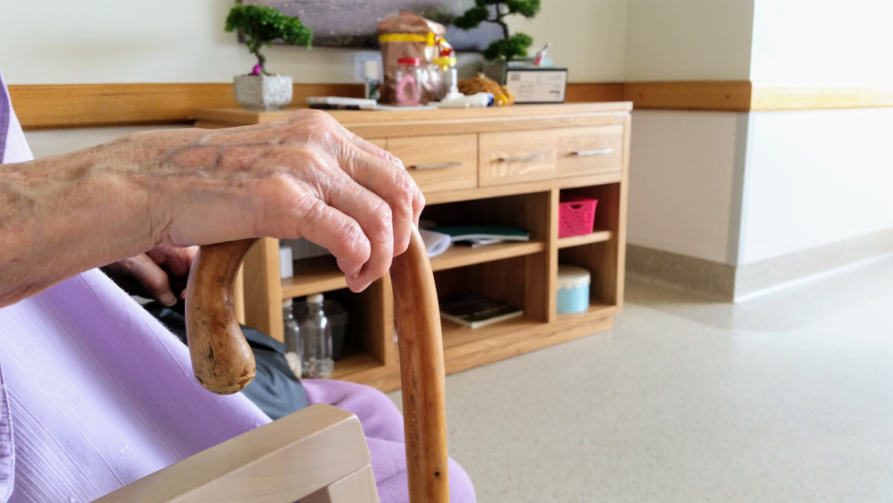 The image shows an elderly person's hand resting on a wooden cane.