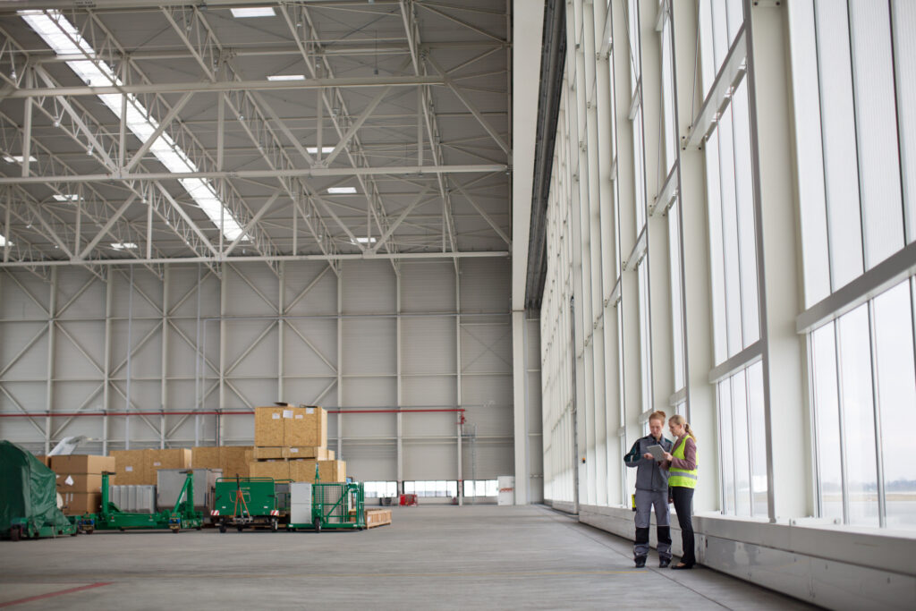 Two individuals standing in an empty warehouse, surrounded by bare walls and a concrete floor.